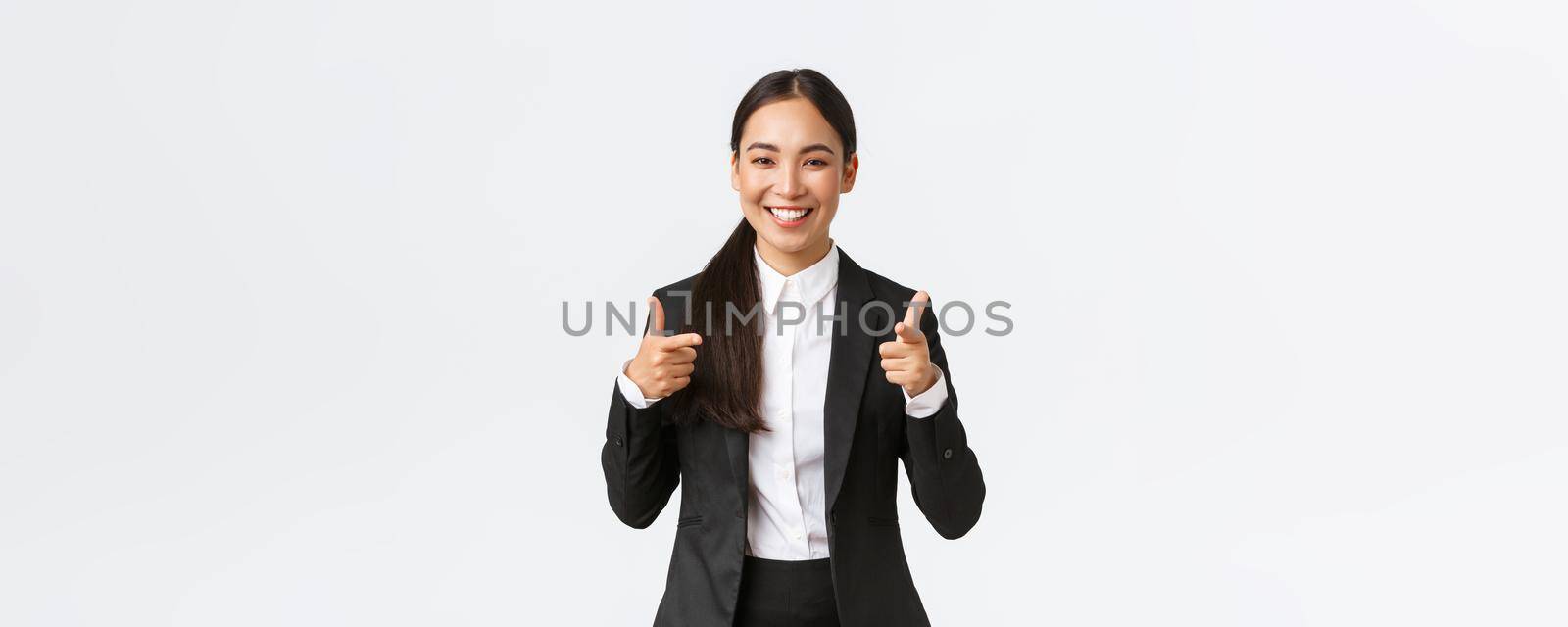 Confident smiling asian saleswoman in black suit showing thumbs-up, guarantee quality of product or providing best service. Sales manager trying sell something to client, white background.