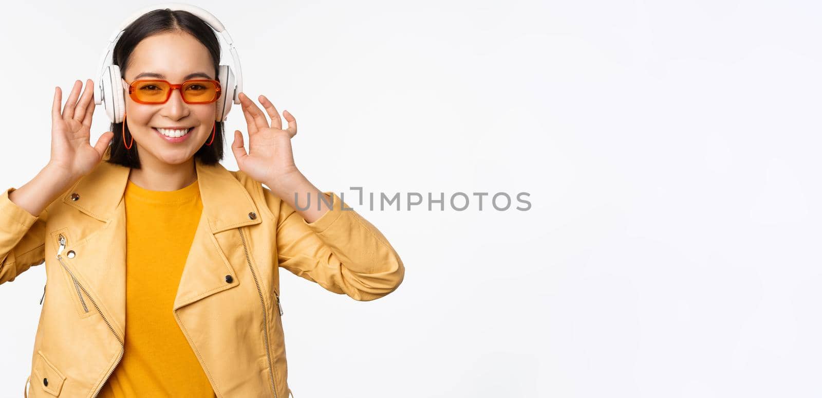 Beautiful asian girl, laughing happy, listening music in headphones, standing over white background by Benzoix