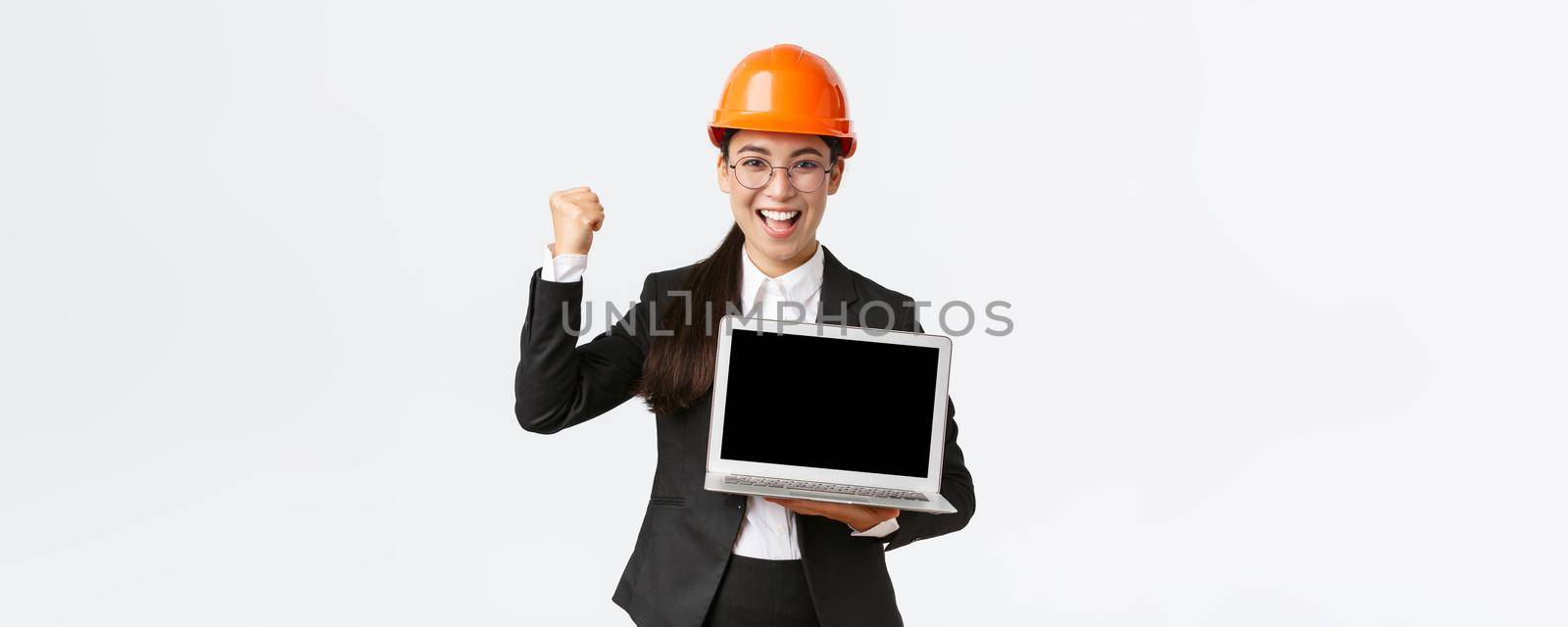 Successful winning businesswoman, construction manager or engineer at factory showing profit chart, diagram at laptop screen, fist pump in rejoice, standing pleased over white background by Benzoix