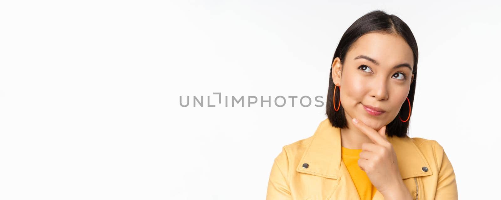 Close up of asian woman thinking, looking up thoughtful, pondering smth with pleased smiling face, standing over white background.