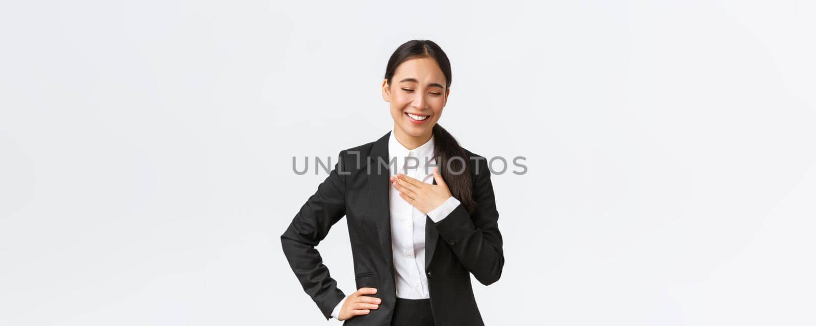 Professional successful businesswoman in black suit having conversation, laughing and touching chest as her something funny or flattering, being praised for good job at work, white background.