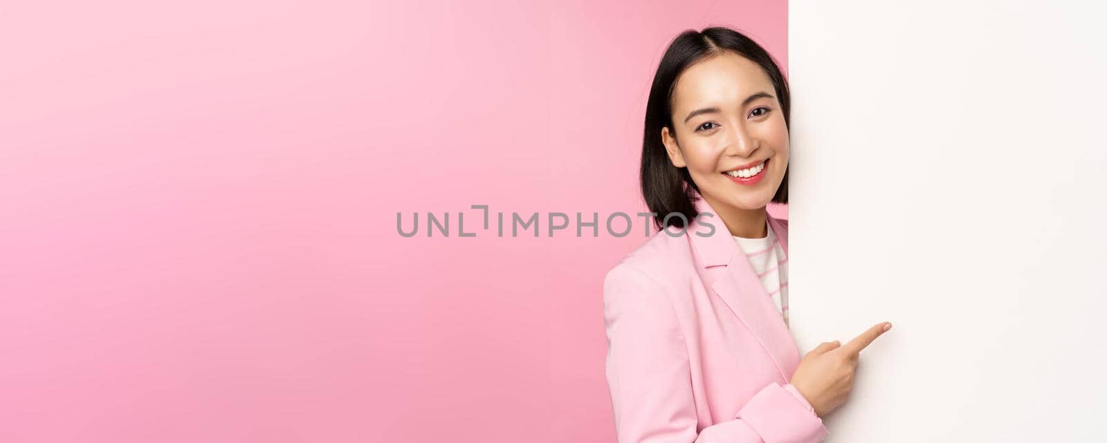 Image of asian female entrepreneur in suit, pointing finger at board, showing smth on white wall, demonstrating chart or information, standing over pink studio background.