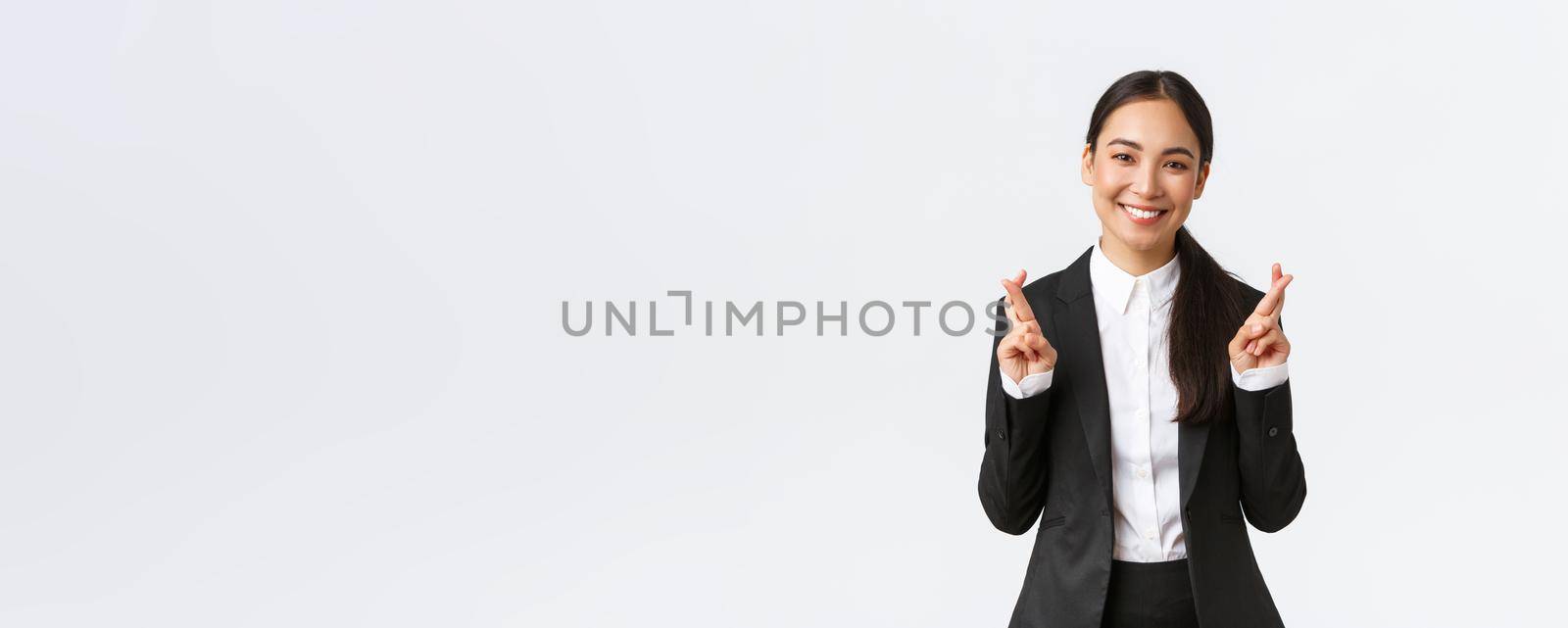 Hopeful businesswoman awaiting for promising results, cross fingers good luck and smiling optimistic. Happy saleswoman in black suit making wish, anticipating good news, white background by Benzoix