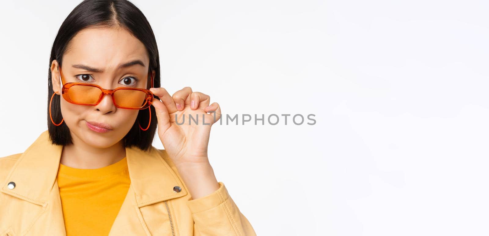 Image of young asian stylish woman, looking from under sunglasses with skeptical, disbelief face, express doubt, standing over white background by Benzoix