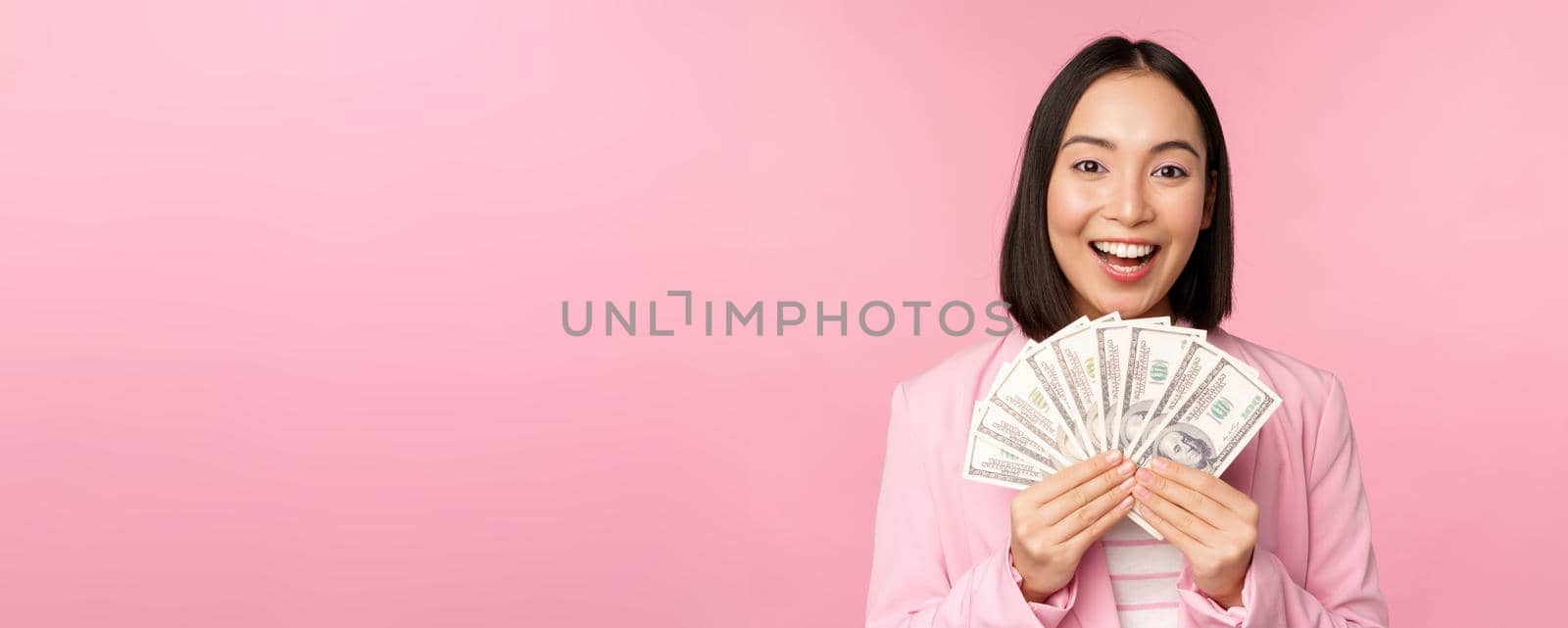 Finance, microcredit and people concept. Happy smiling asian businesswoman showing dollars money, standing in suit against pink background by Benzoix