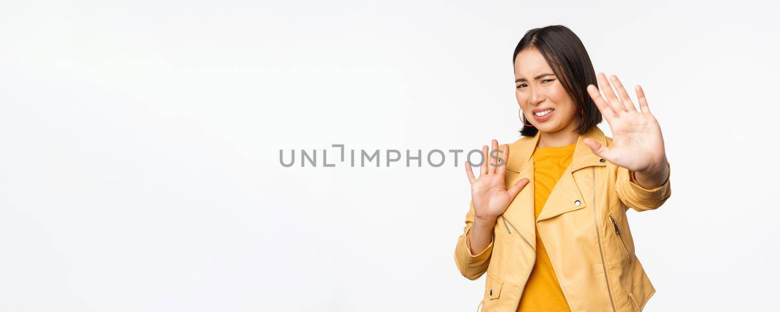 Stop, get away from me, back off concept. Young disgusted asian woman being blinded by smth, stretching out hands in defense, blocking light, standing over white background by Benzoix