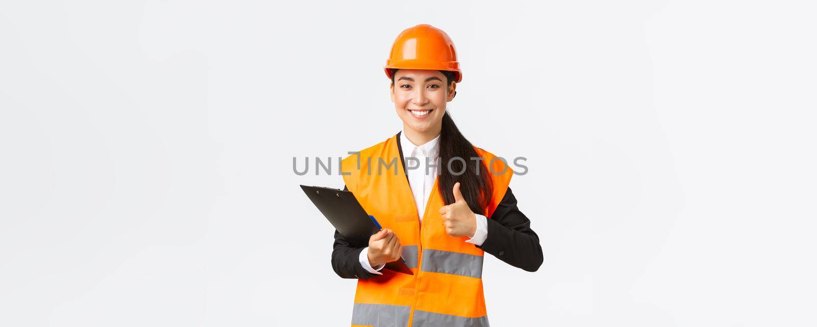 Satisfied asian female construction engineer pleased with results of inspection, wearing safety clothing and helmet at building area, writing down notes and show thumbs-up in approval.