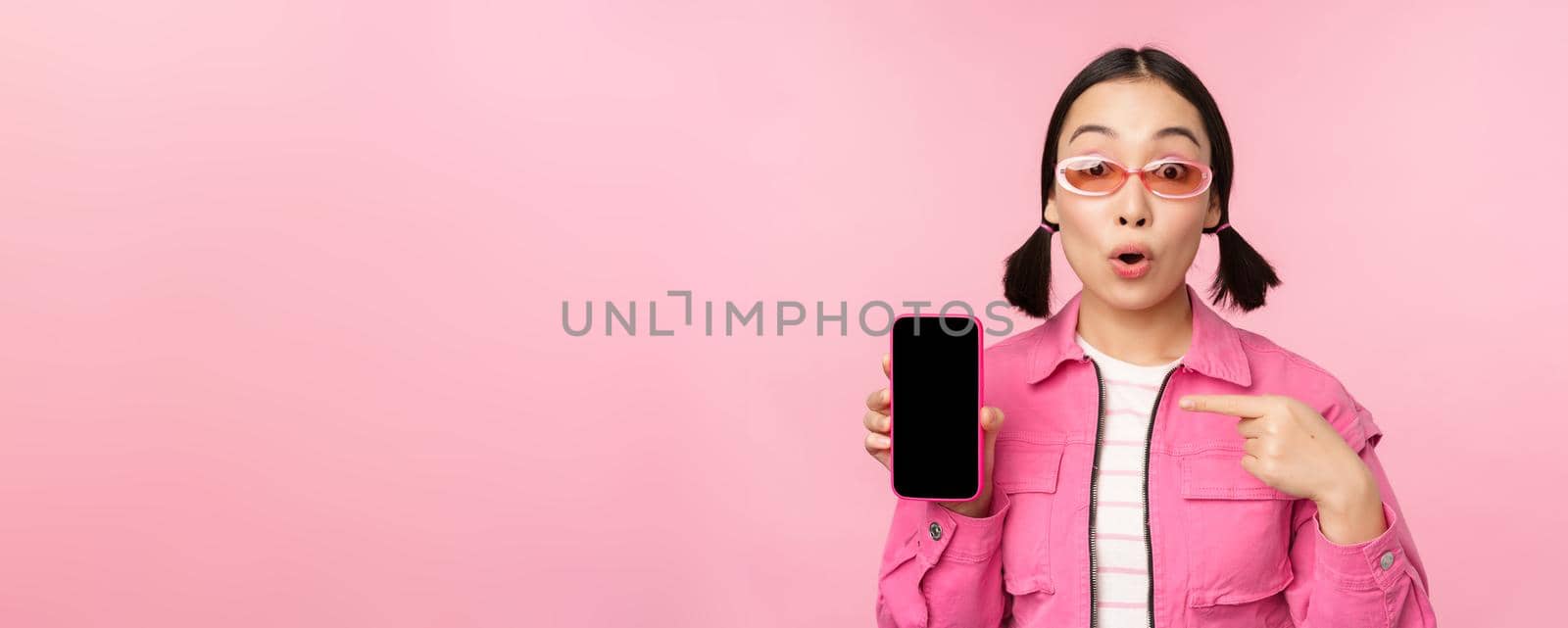 Enthusiastic asian woman in stylish clothes, sunglasses, pointing finger at mobile phone screen, showing smartphone application, standing over pink background.