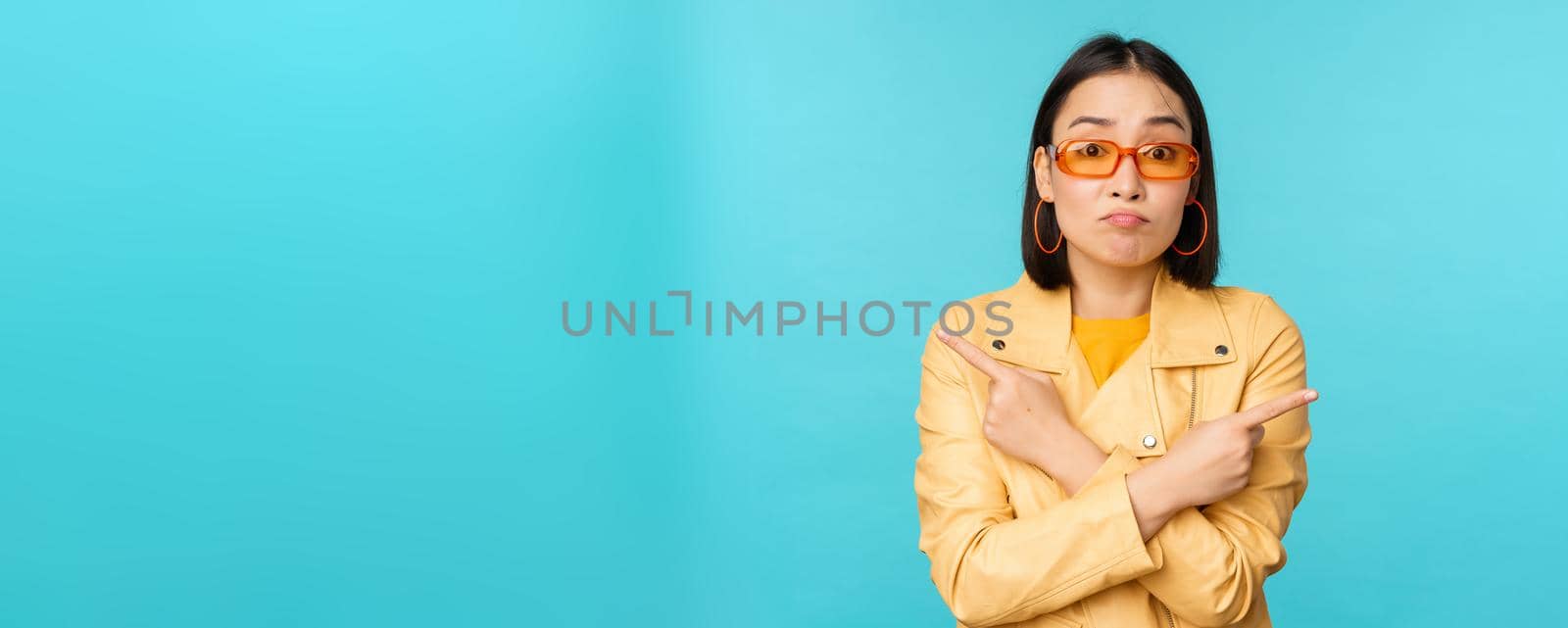 Indecisive asian woman in sunglasses, makes choice between two variants, points sideways, looks puzzled, stands over blue background by Benzoix