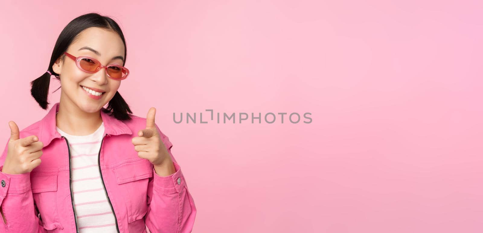 Close up portrait of modern asian girl in sunglasses smiling, pointing fingers at camera, praise you, inviting or complimenting, standing over pink background.