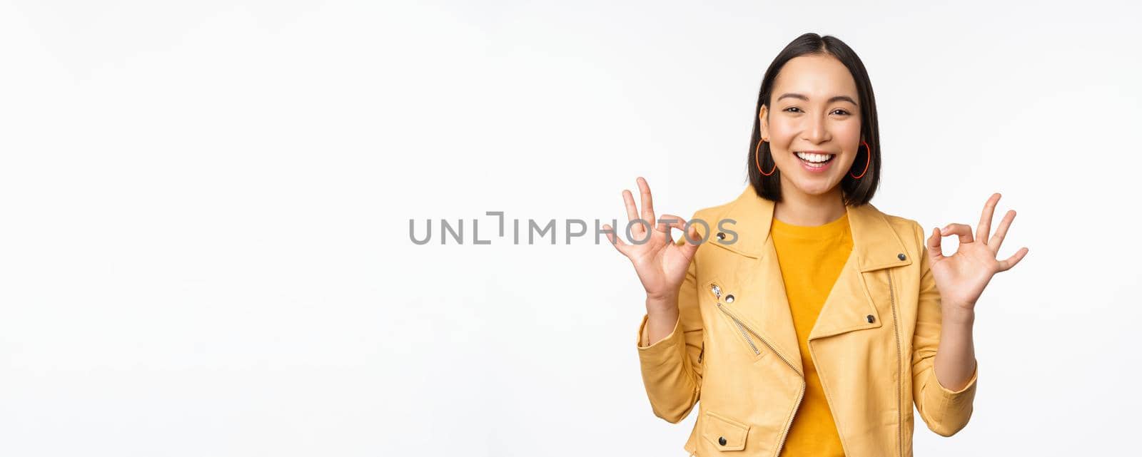 Happy beautiful asian girl shows okay, ok recommending sign, pleased by smth, praising quality, standing over white background. Copy space