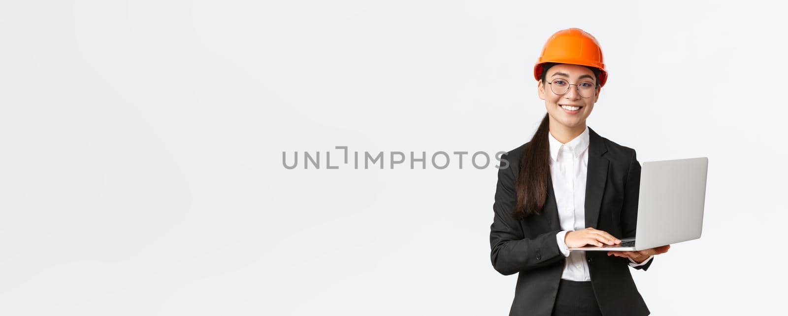 Smiling professional female saleswoman showing construction works on new house. Asian industrial engineer in safety helmet and business suit using laptop computer, enter data from factory by Benzoix