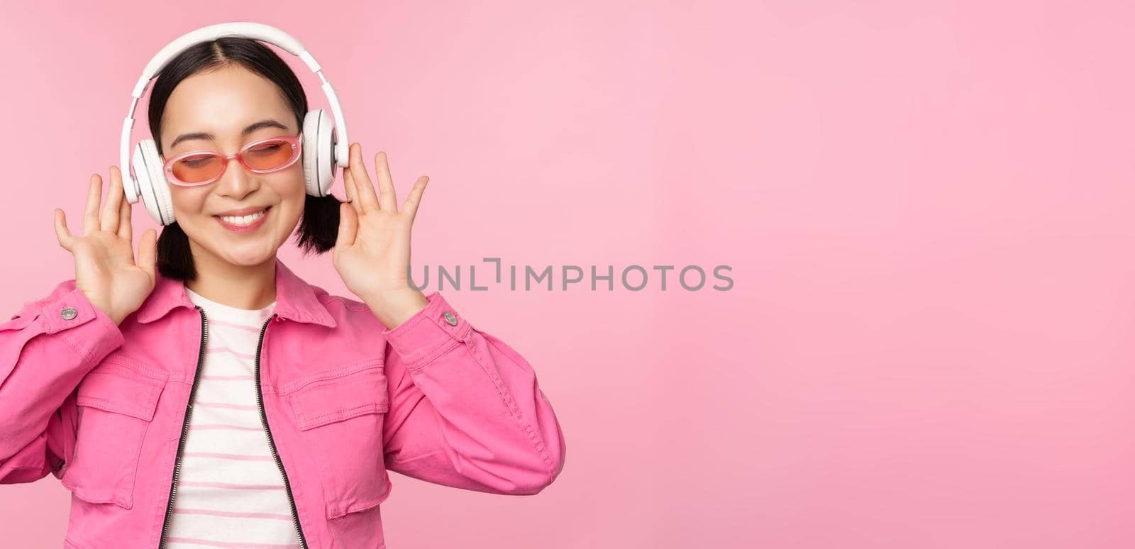 Dancing stylish asian girl listening music in headphones, posing against pink background by Benzoix