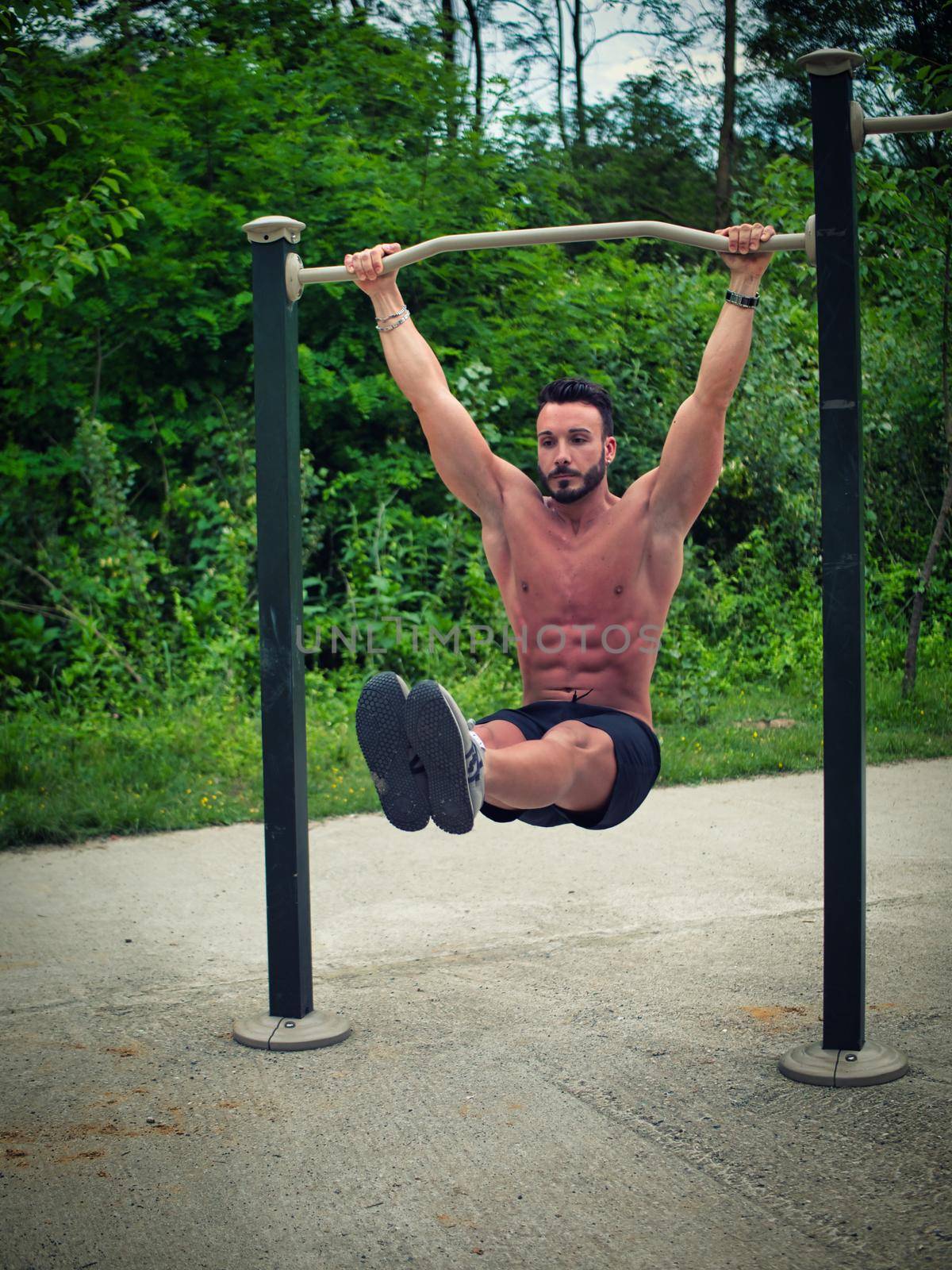 Attractive shirtless young man hanging from bar and working out in outdoor gym in city park