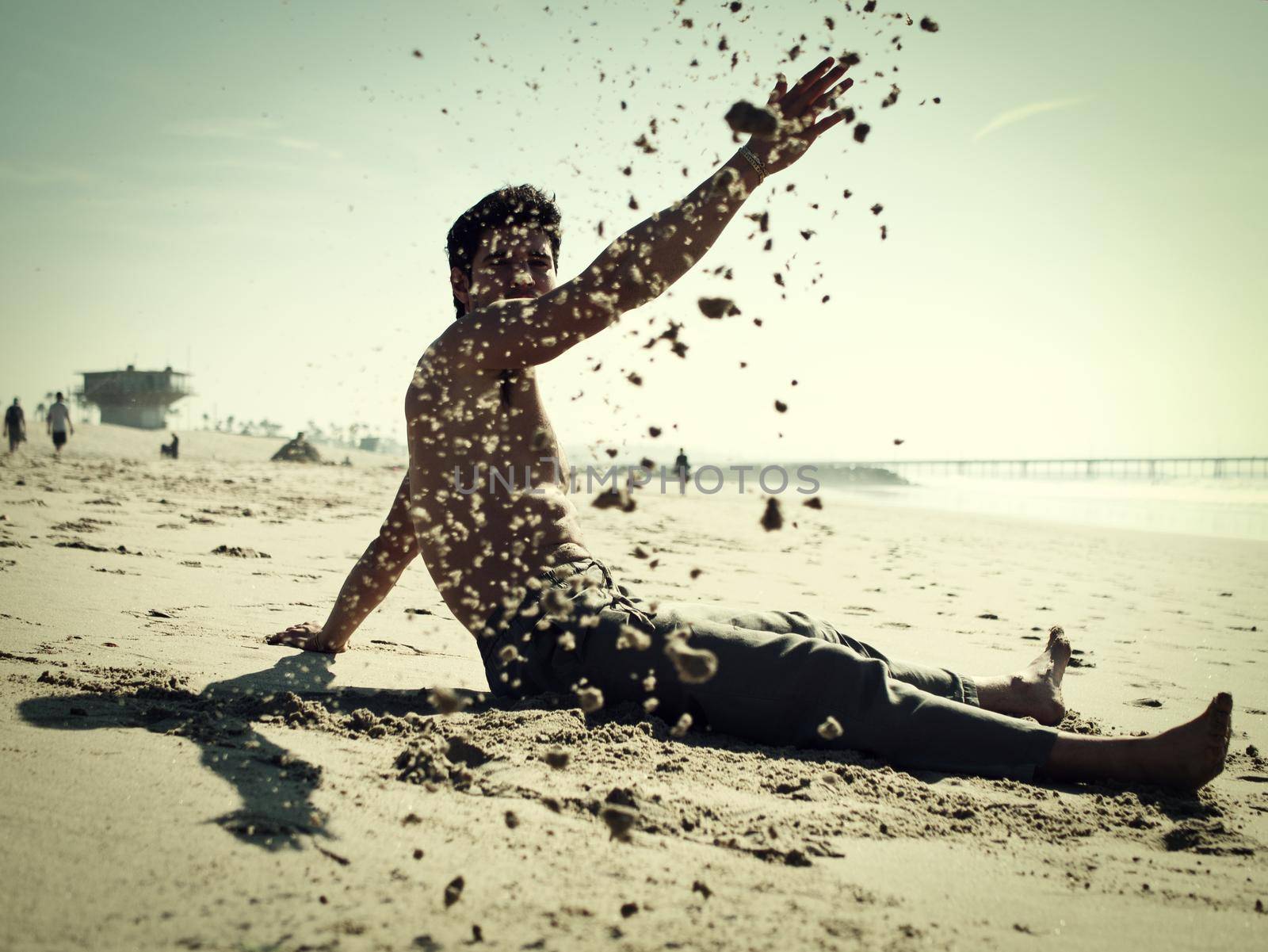 Handsome young man sitting on a beach playing with sand, shirtless wearing boxer shorts, showing muscular fit body