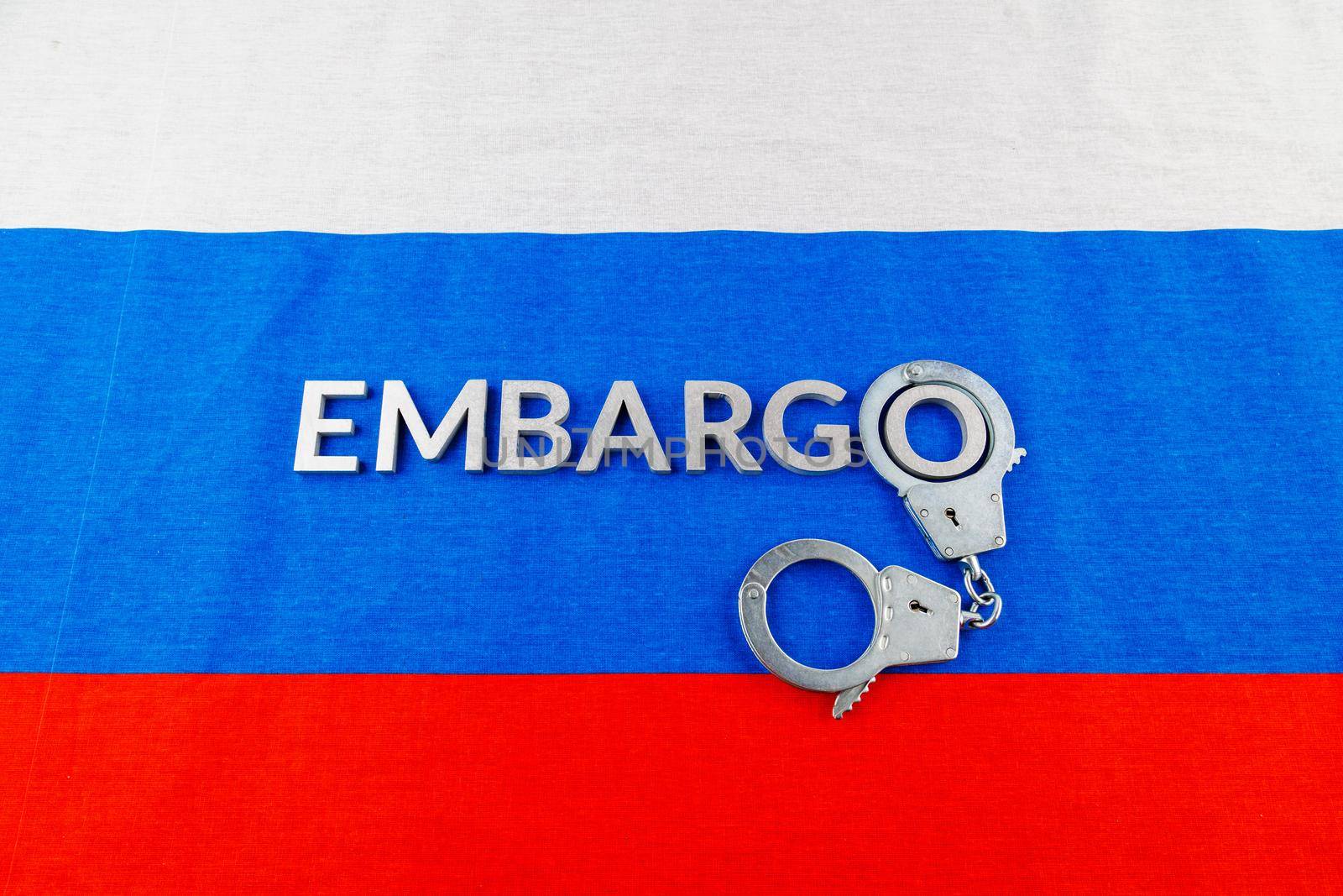 the word embargo laid with silver metal letters on russian tricolor flag with silver handcuffs in linear perspective