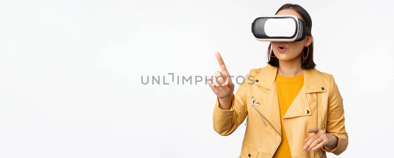 Young asian girl wearing VR glasses, looking amazed, using virtual reality headset, posing against white background by Benzoix