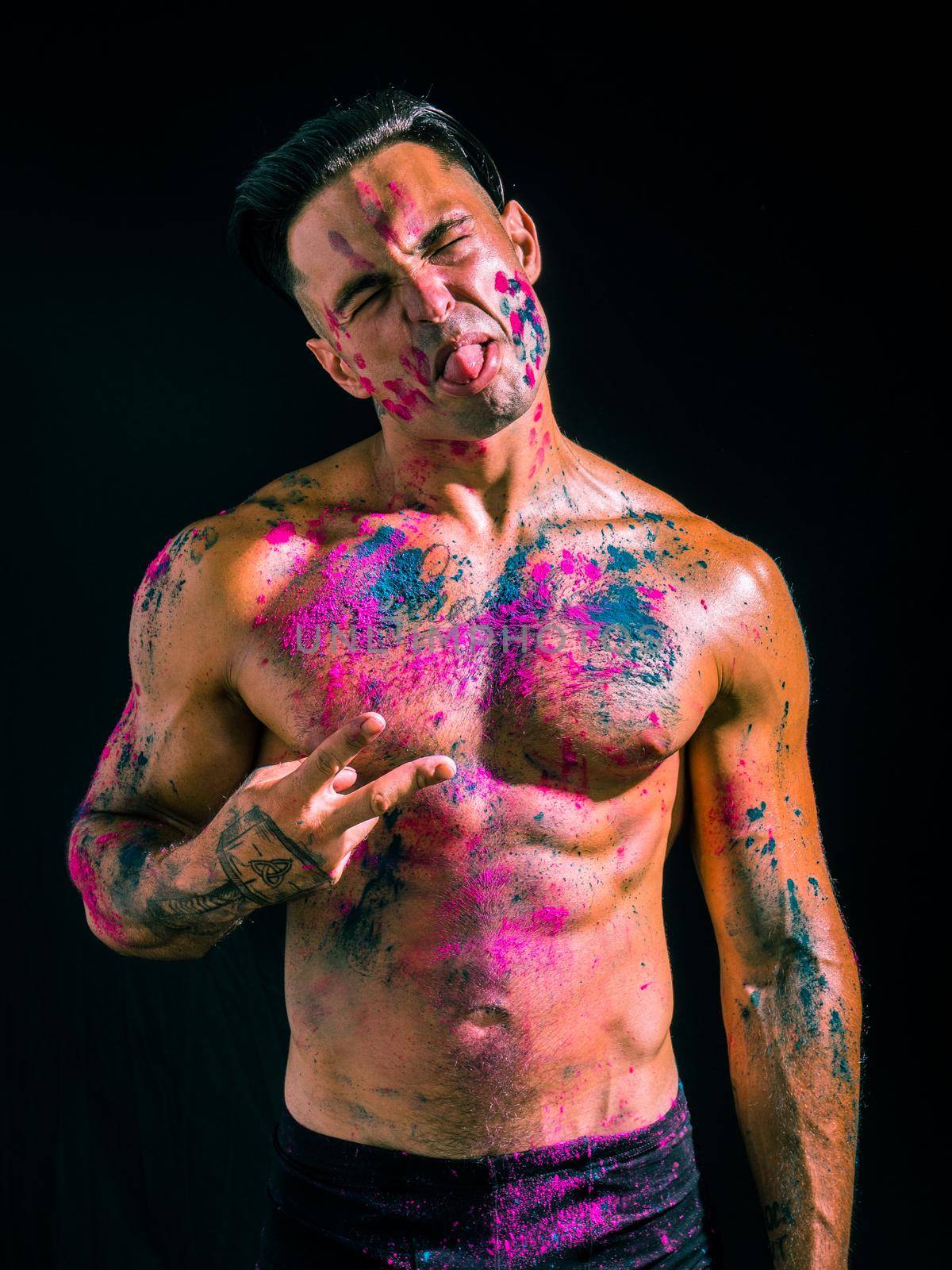 Muscular young man shirtless with skin painted with Holi colors, looking at camera with tongue sticking out