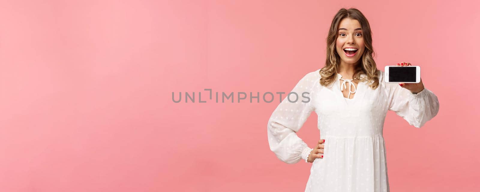 Portrait of cheerful, upbeat attractive blond caucasian girl in white dress, showing smartphone display, hold mobile phone horizontal smiling camera amazed, stand pink background.