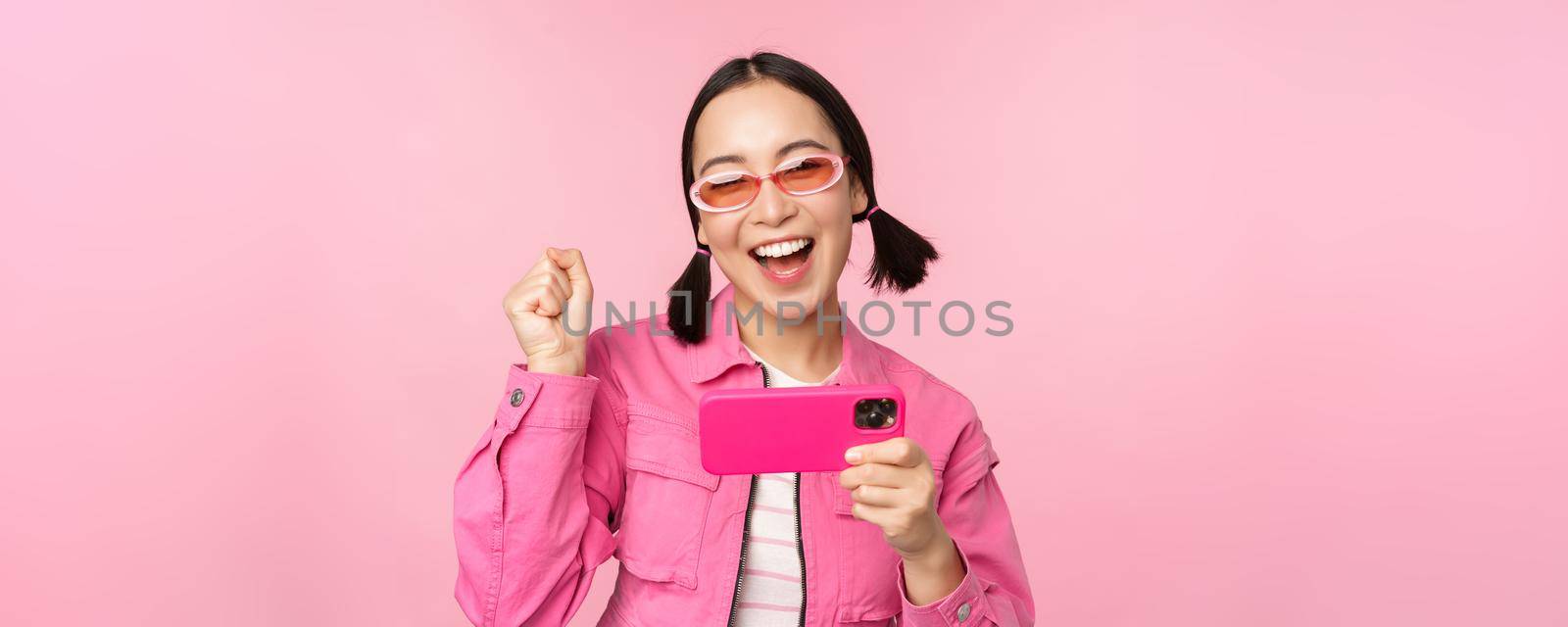 Happy smiling korean girl winning on mobile phone, looking at horizontal smartphone screen and rejoicing, achieve goal, celebrating, standing over pink background.
