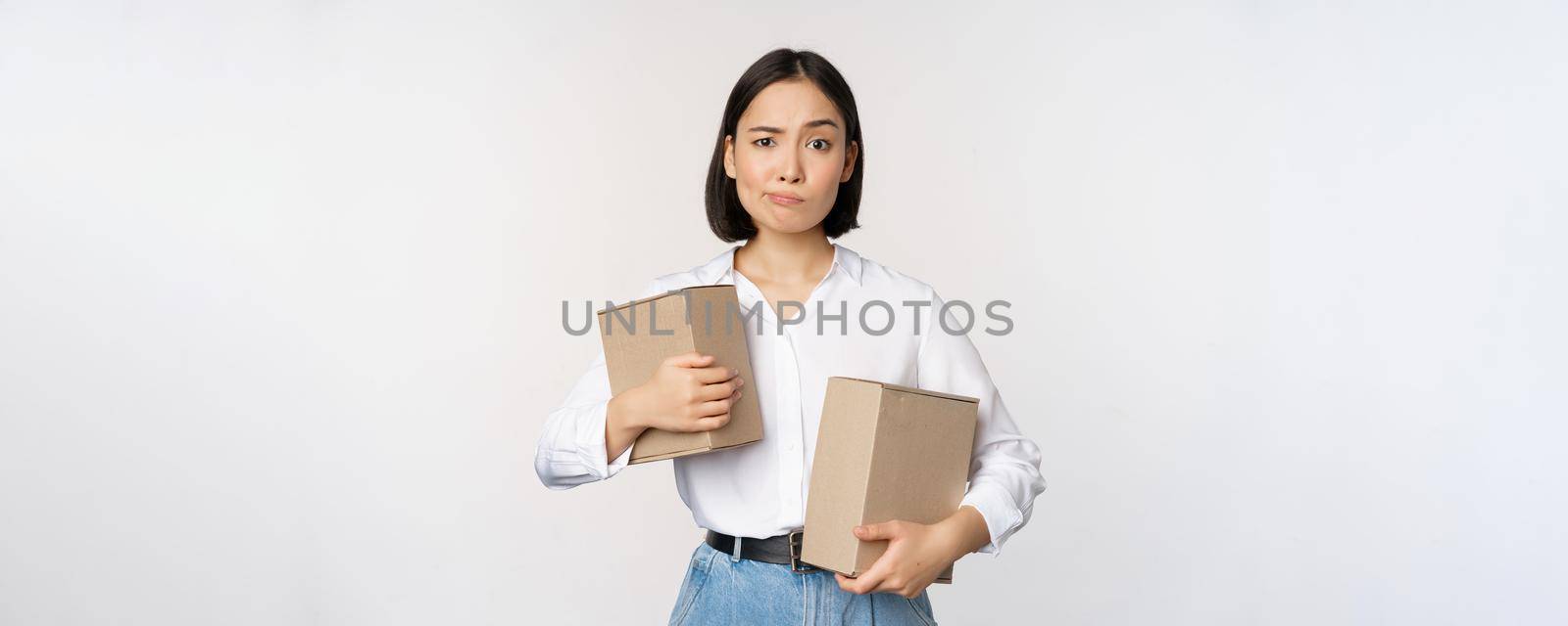 Complicated young asian woman holding two boxes, looking doubtful at camera, standing over white background puzzled by Benzoix