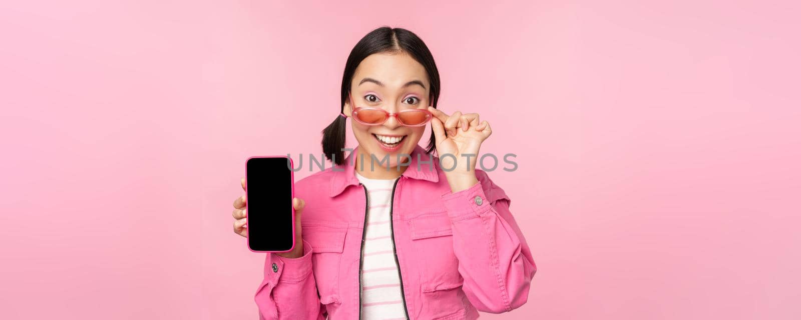 Portrait of asian girl showing mobile phone screen, reacting surprised, standing over pink background by Benzoix