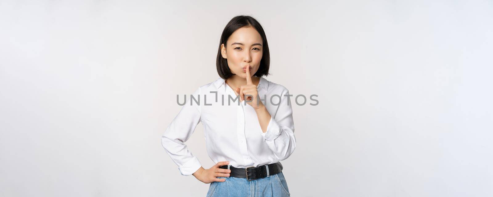 Portrait of young asian adult woman shushing, say hush shh, press finger to lips, sharing secret, dont speak taboo gesture, standing over white background.