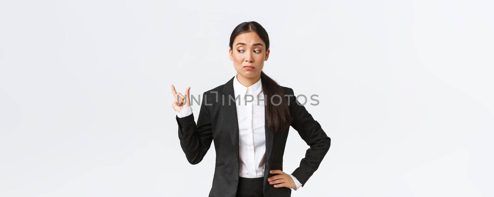 Skeptical and awkward young asian businesswoman, saleswoman in black suit shaping something small and looking disappointed in size, grimacing unamused over little tiny thing, white background.