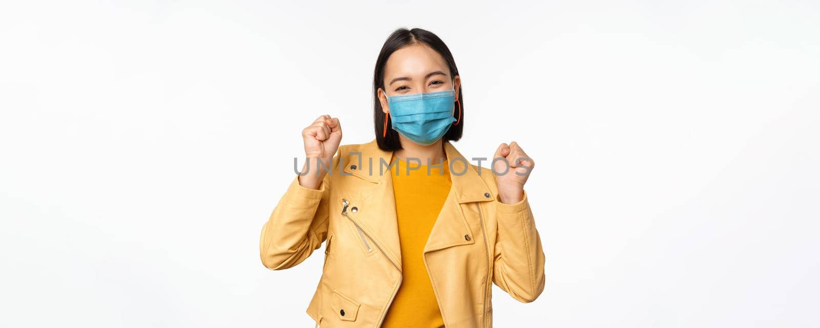 Covid-19 and people concept. Young asian woman in medical face mask, dancing happy in casual clothes, posing over white background by Benzoix