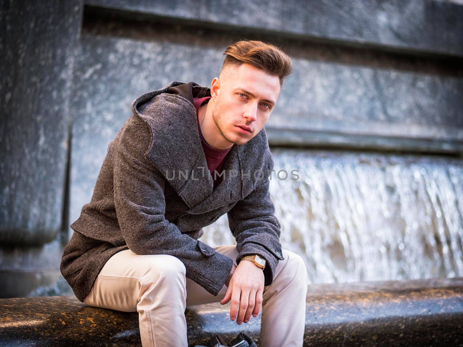 Young man, a student, sitting by fountain, looking at camera in city setting in a winter day