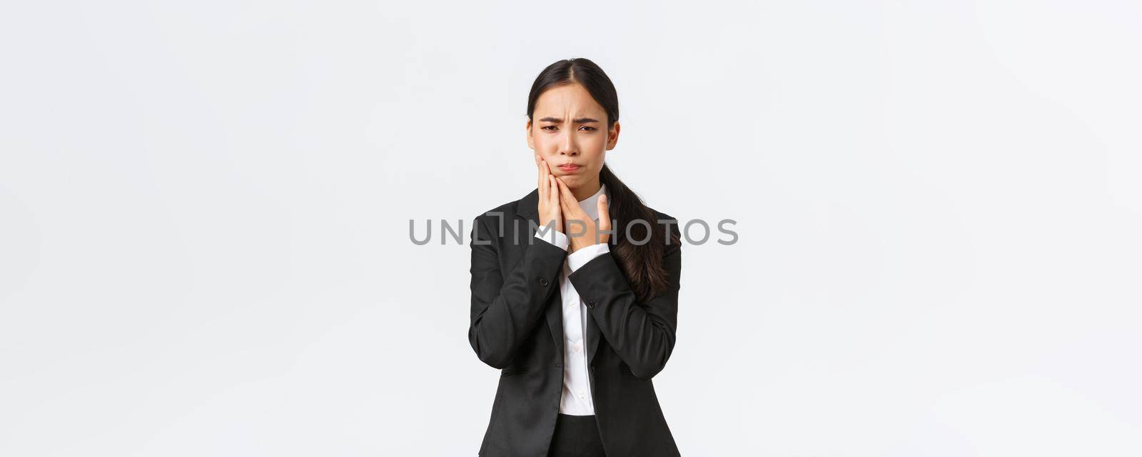 Female asian office manager in black suit having toothache at work. Troubled businesswoman holding hand on cheek as feeling pain in teeth, need appointment doctor, standing white background.
