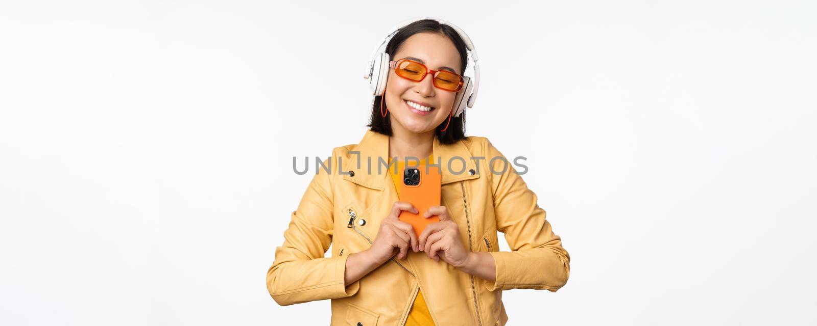 Technology concept. Stylish asian girl in headphones, holding smartphone, dancing and singing, listening music, standing over white background by Benzoix