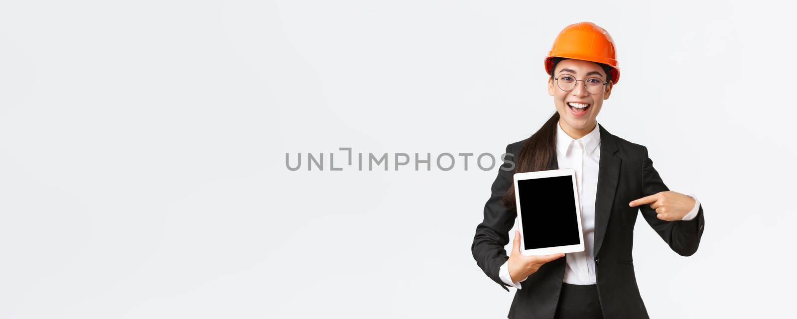 Smiling professional female construction manager, asian engineer in safety helmet and business suit show project, pointing finger at digital tablet screen with pleased expression, white background.