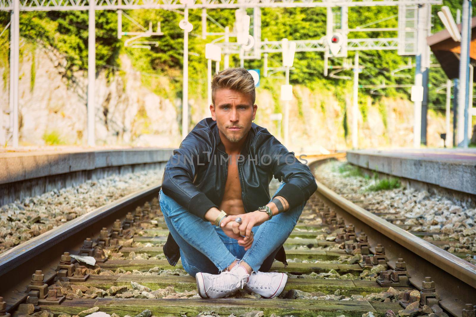 Attractive young man sitting on railroad, wearing black leather jacket, sneakers and jeans, looking at camera, at daytime