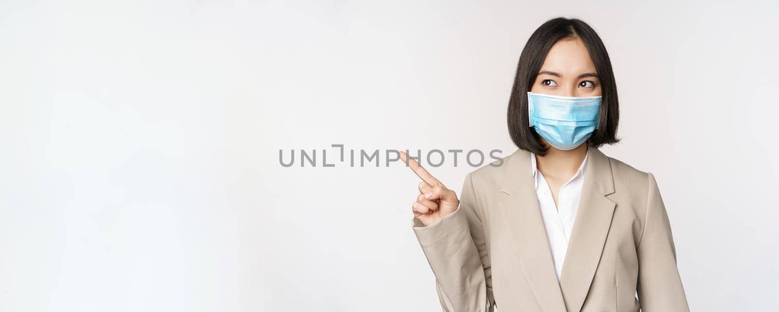 Enthusiastic businesswoman pointing fingers left, wearing medical face mask from covid-19 pandemic, standing over white background by Benzoix