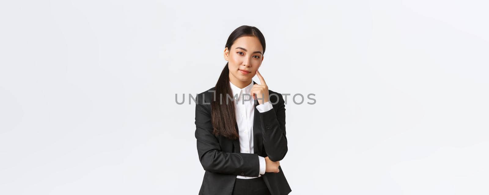 Creative smart asian female manager, saleswoman in suit smiling cunning and looking at camera, have plan, thinking, got excellent idea, standing pleased over white background.