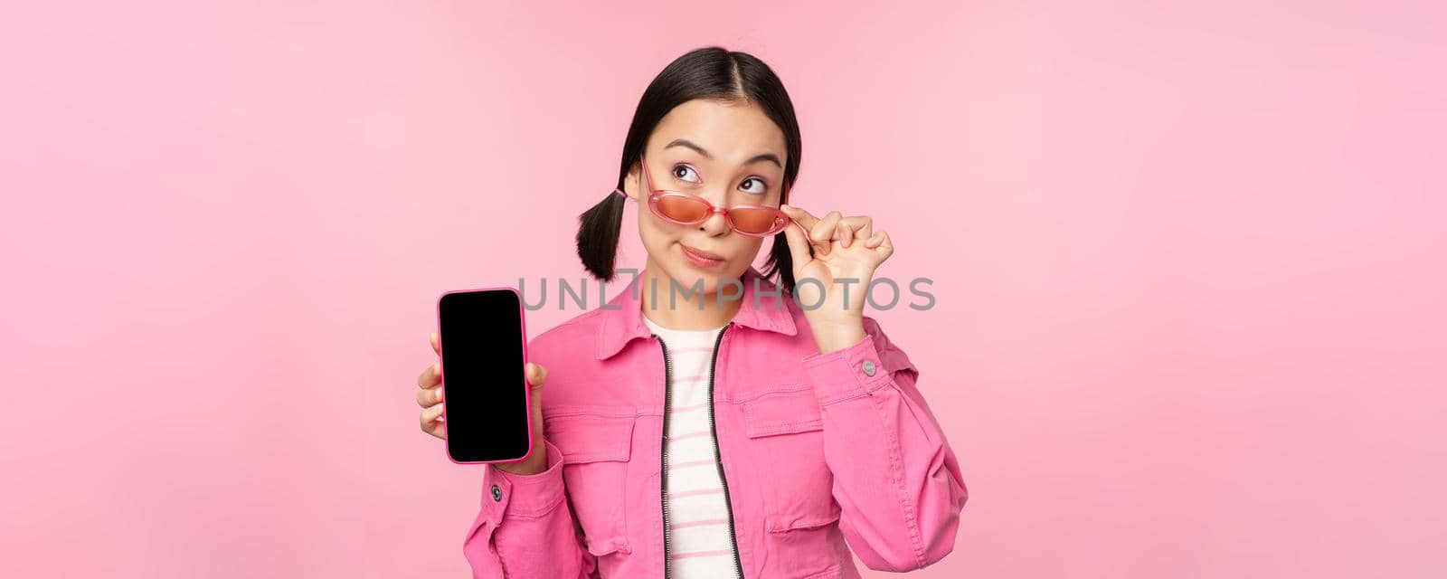 Stylish korean girl, young woman in sunglasses showing smartphone screen, mobile phone app interface or website, standing over pink background by Benzoix