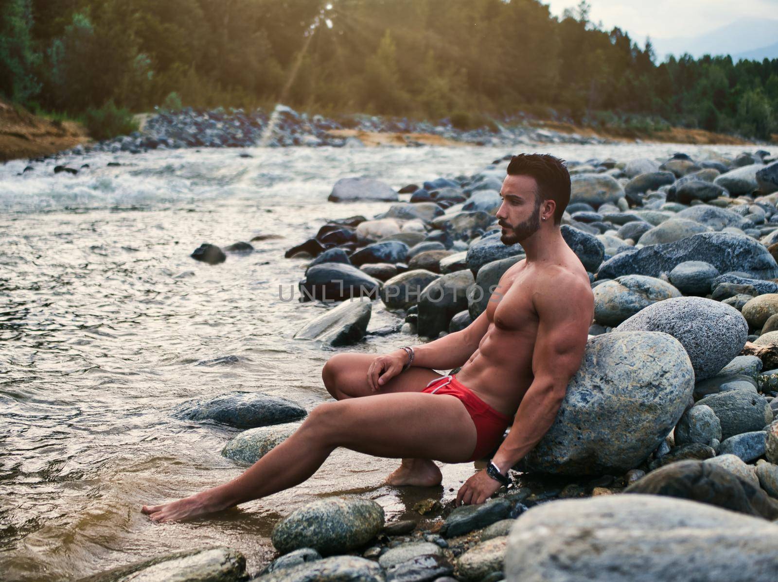 Attractive muscular shirtless young man in nature next to a river, wearing swimming suit, sitting on rock