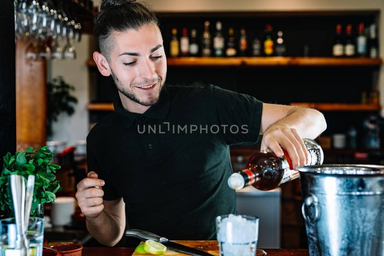 Young and modern waiter, with long dark hair, dressed in black polo shirt, adding liquor in a crystal glass full of ice to prepare a Mojito. Waiter preparing a cocktail. Cocktail glass with ice cubes. Mojito. Bar full of cocktail ingredients. Dark background and dramatic lighting. Horizontal