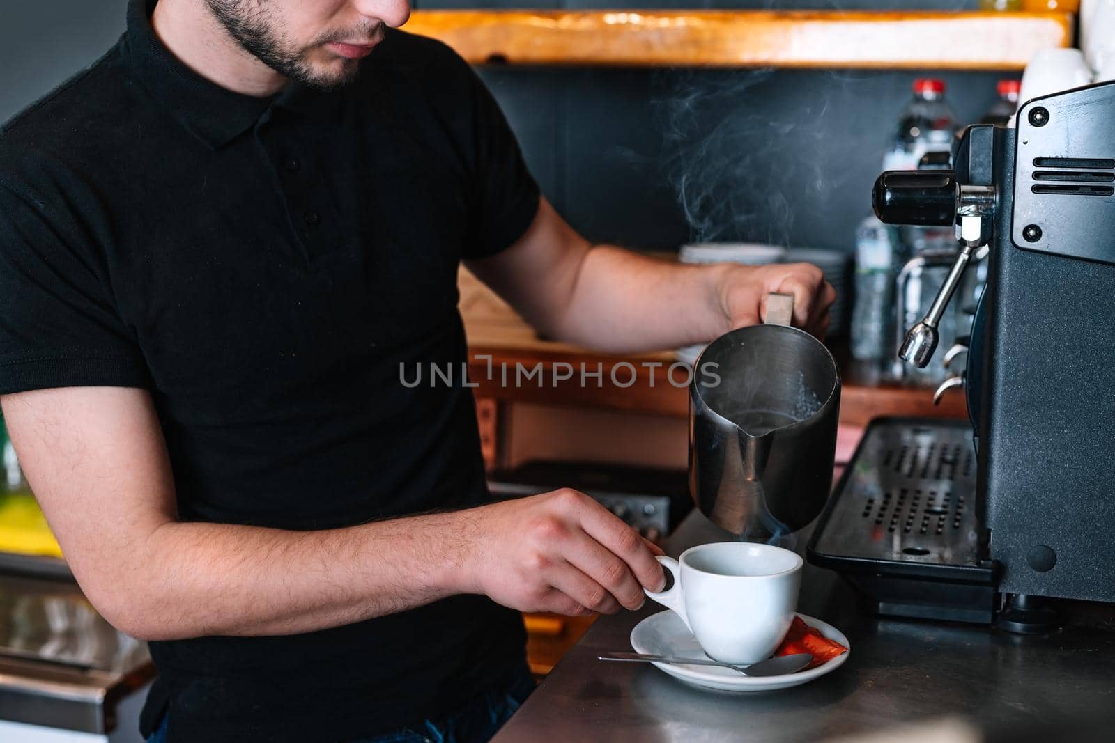 detail of the hands of a young waiter, concentrated and hard-working, dressed in the company uniform, black polo shirt, serving coffee in a white cup. Restaurant. Preparing a coffee for a customer. Preparing coffee with milk. Warm atmosphere and dim lighting. Steaming cups of coffee. Horizontal