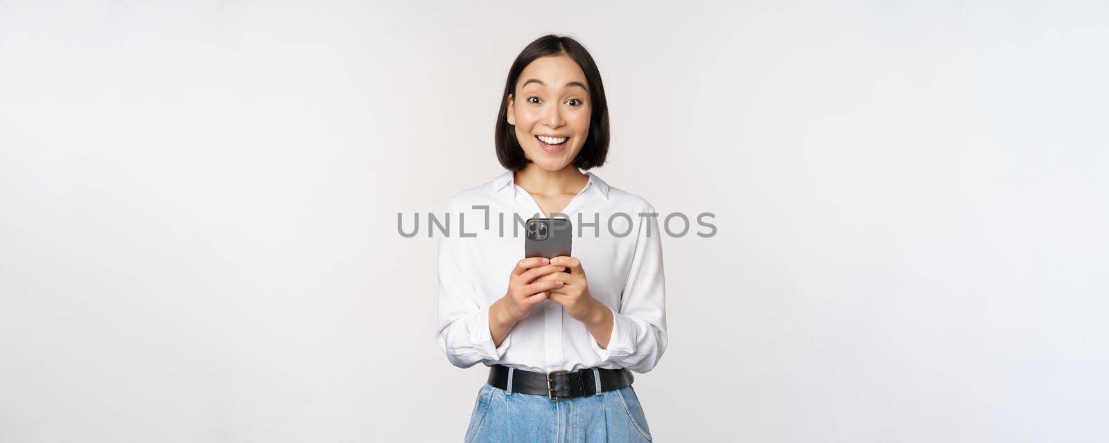 Excited asian woman smiling, reacting to info on mobile phone, holding smartphone and looking happy at camera, standing over white background.