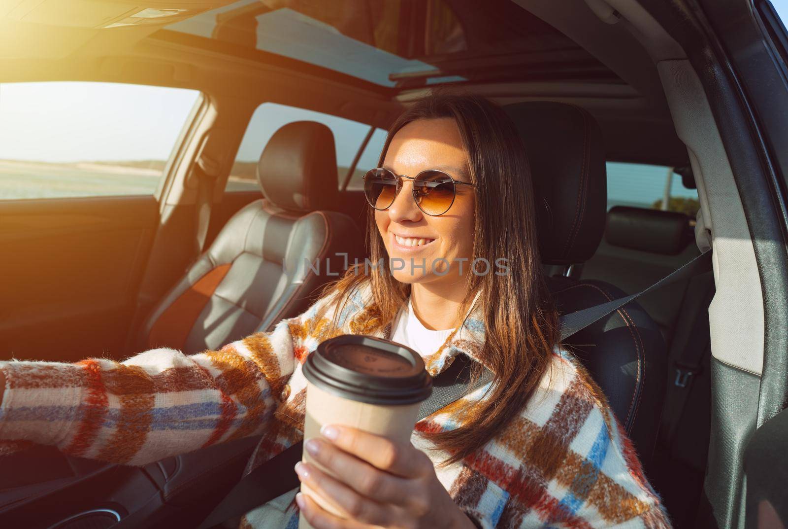 Happy smiling beautiful woman holding coffee cup and driving a car on a sunny day by DariaKulkova