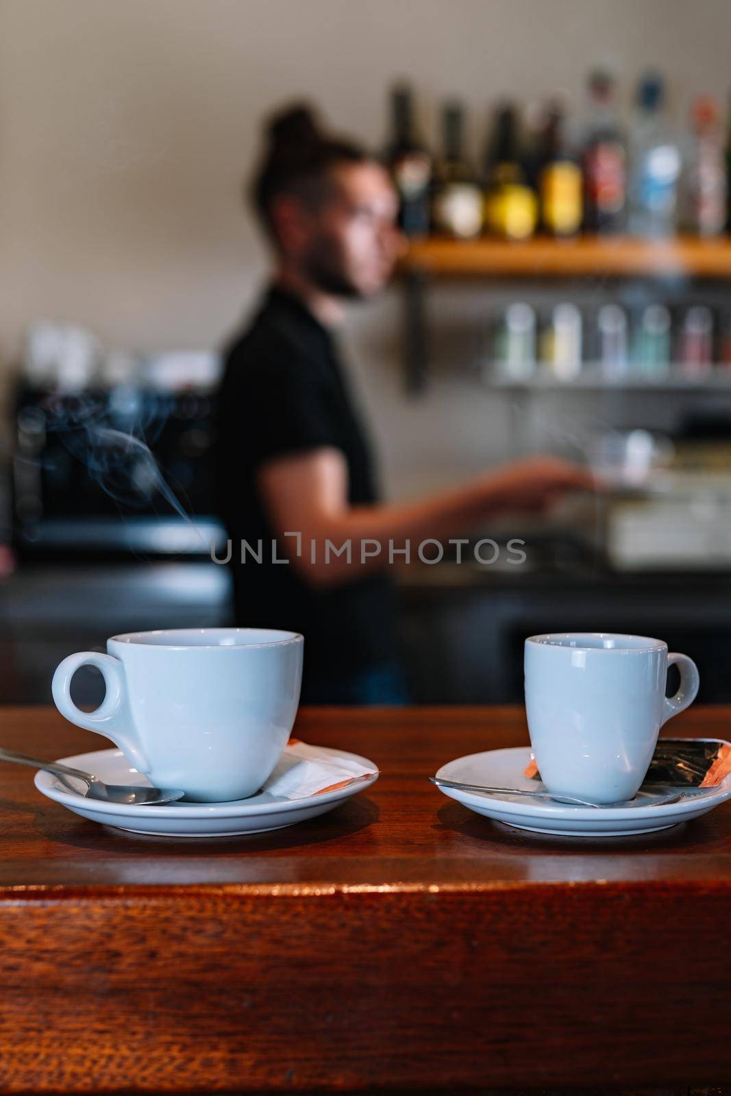 Detail shot of two white coffee cups on the bar counter. Waiting to be served to the customers. Background young waiter in black T-shirt. Warm atmosphere and dim light. Steaming cups of coffee. Vertical