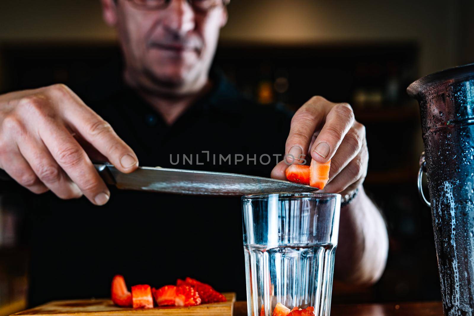 Detail of the hands of an experienced waiter, concentrated and precise, dressed in the company uniform, black polo shirt, pouring cut strawberries into a crystal glass on the counter of the nightclub. Preparing cocktails for customers. Warm atmosphere and dim light. Horizontal