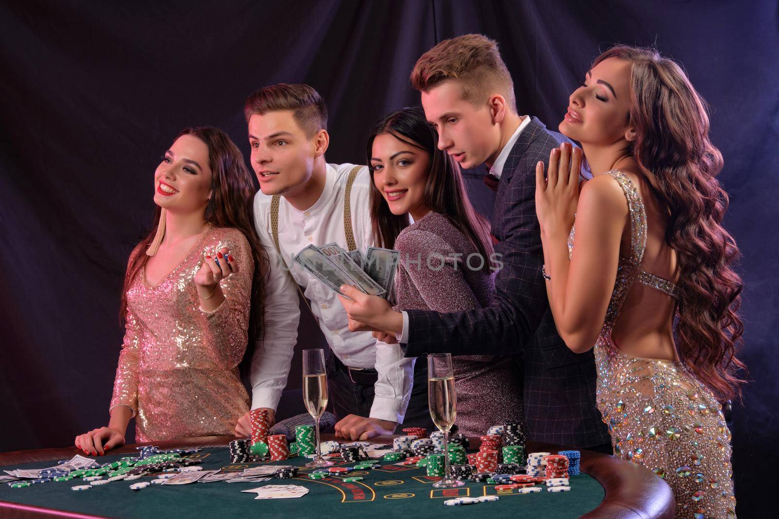 Friends are playing poker at casino, at table with stacks of chips, money, cards, champagne on it. Celebrating their win, smiling. Black background. Gambling entertainment. Side view. Close-up.