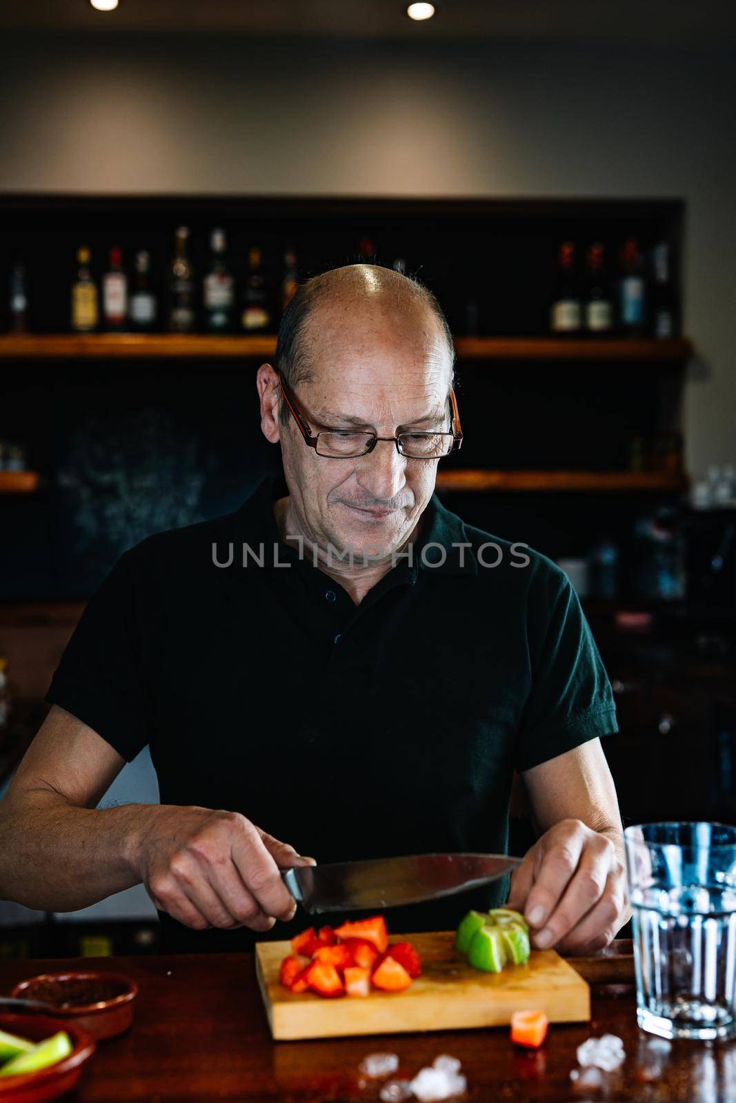 Experienced waiter, concentrated and precise, dressed in company uniform, black polo shirt, pouring cut strawberries into a glass tumbler on the nightclub counter. Preparing cocktails for customers. Warm atmosphere and dim light. Horizontal