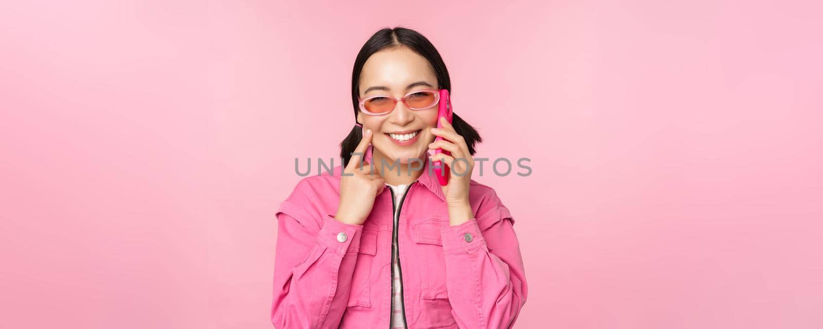 Beautiful korean female model in sunglasses, talking on mobile phone with happy face, using cellular service to call friend on smartphone, standing over pink background by Benzoix