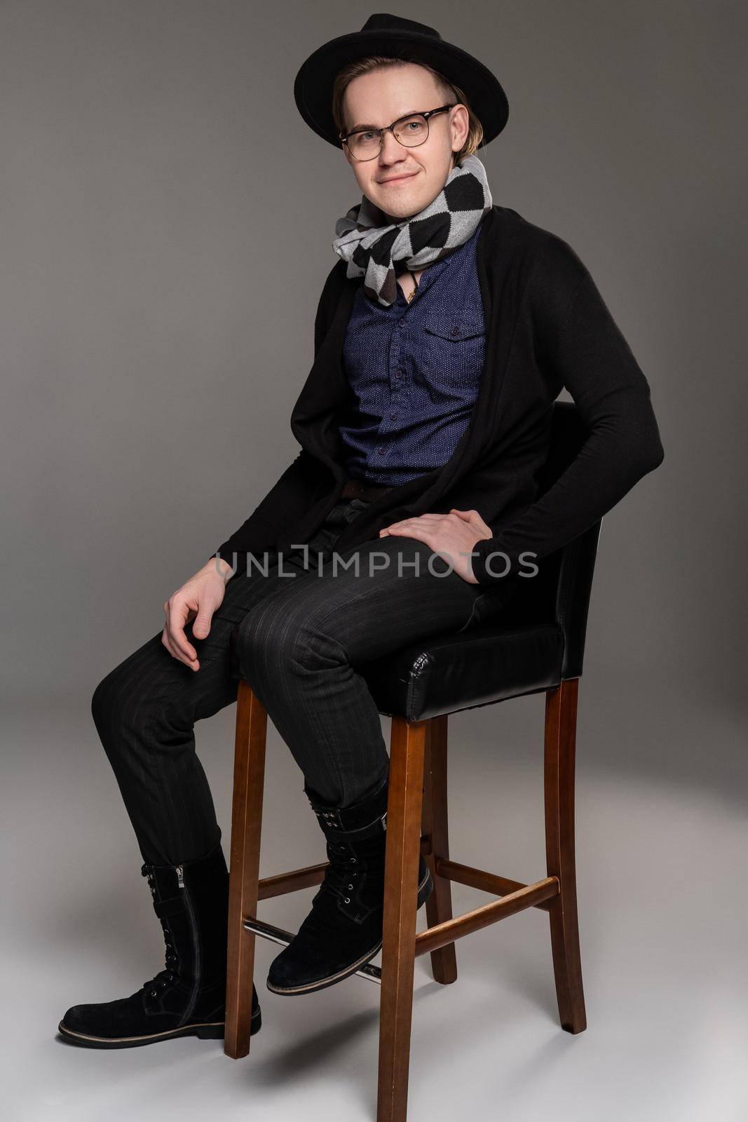 Handsome brunet male in a casual dark clothes, stylish scarf, glases and black hat is sitting on the bar chair and looking at the camera while posing over a gray background. Copy space. Emotions concept.