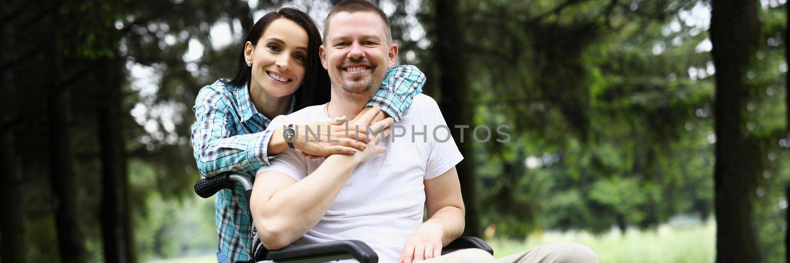 Portrait of happy couple posing in park together on fresh air. Middle aged man sitting in wheelchair, broken leg. Medicine, recovery, love, positive, family and relationship concept