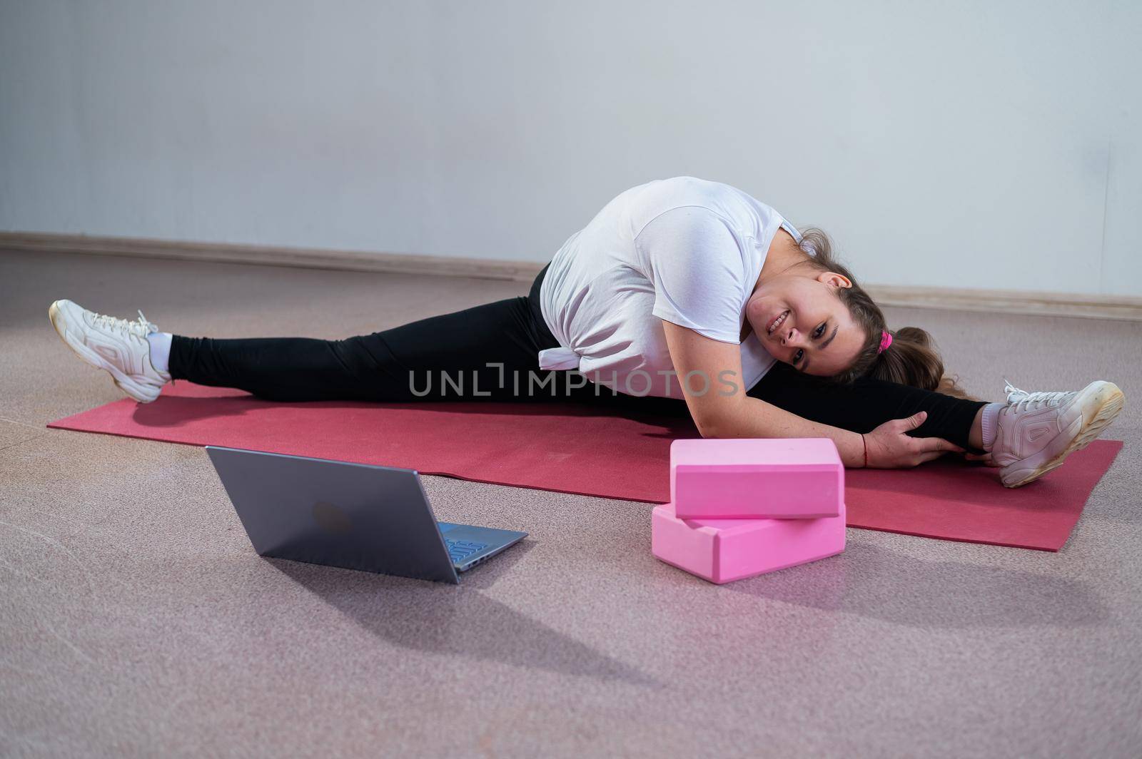 Young caucasian fat woman doing bends on a sports mat and watching a training video on a laptop. A chubby girl stretches the split remotely using video communication by mrwed54